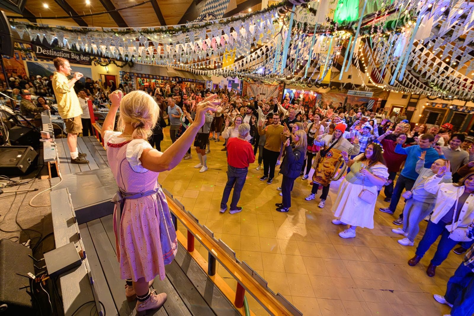 A woman in a dirndl on stage leading a packed dance floor under Bavarian bunting at Big Bear Oktoberfest