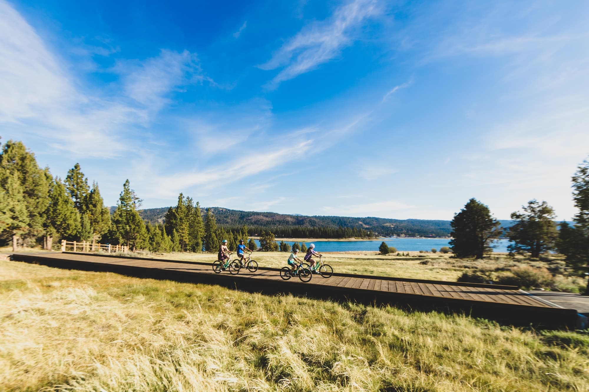 A family riding the Pedal Path Loop's boardwalk through the north-shore meadow, Big Bear Lake behind